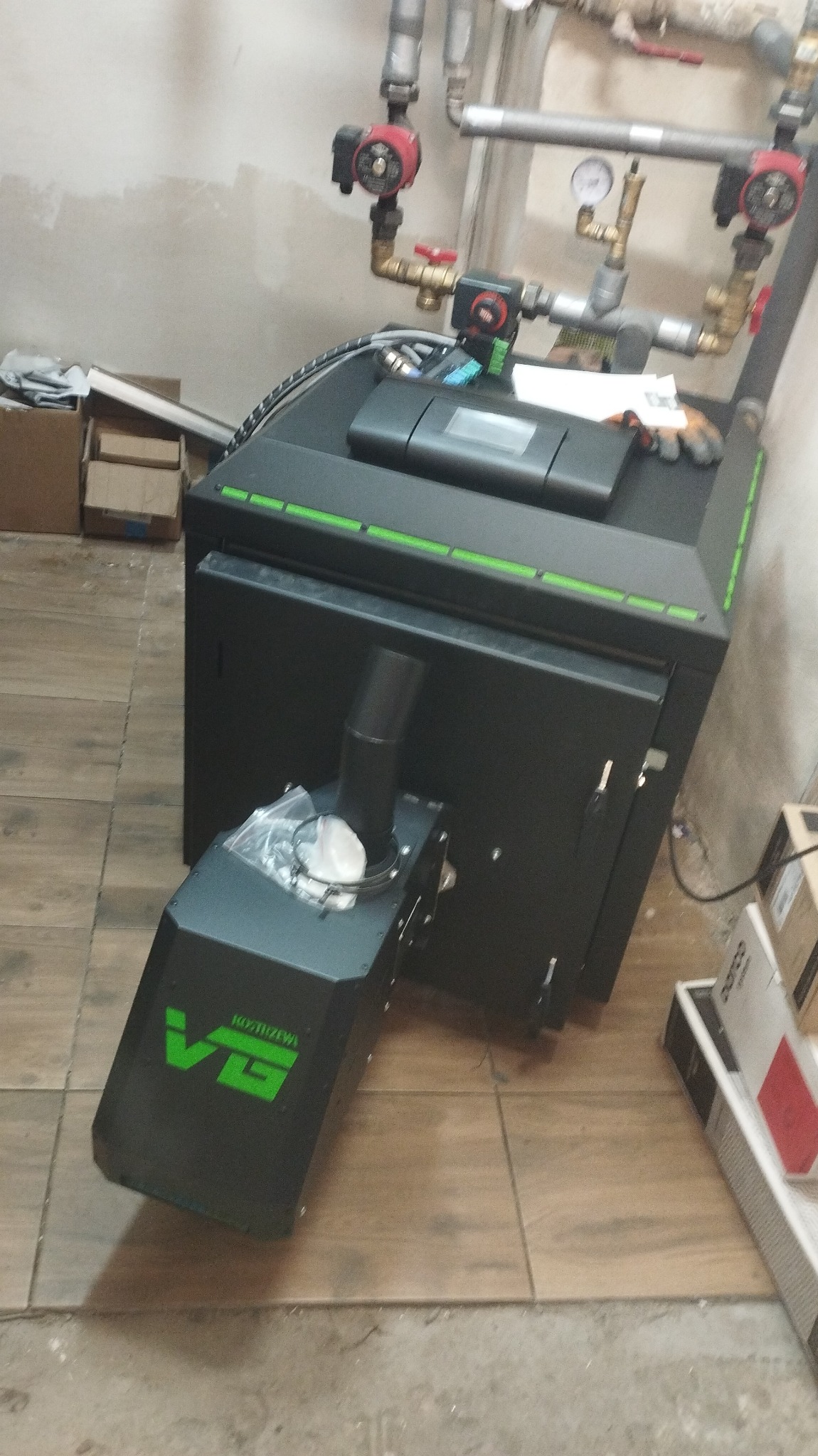 Nowoczesny piec na paliwo stałe, czarny z zielonymi akcentami, z widocznym podajnikiem i elementami instalacji grzewczej w tle, umieszczony na brązowych płytkach podłogowych.