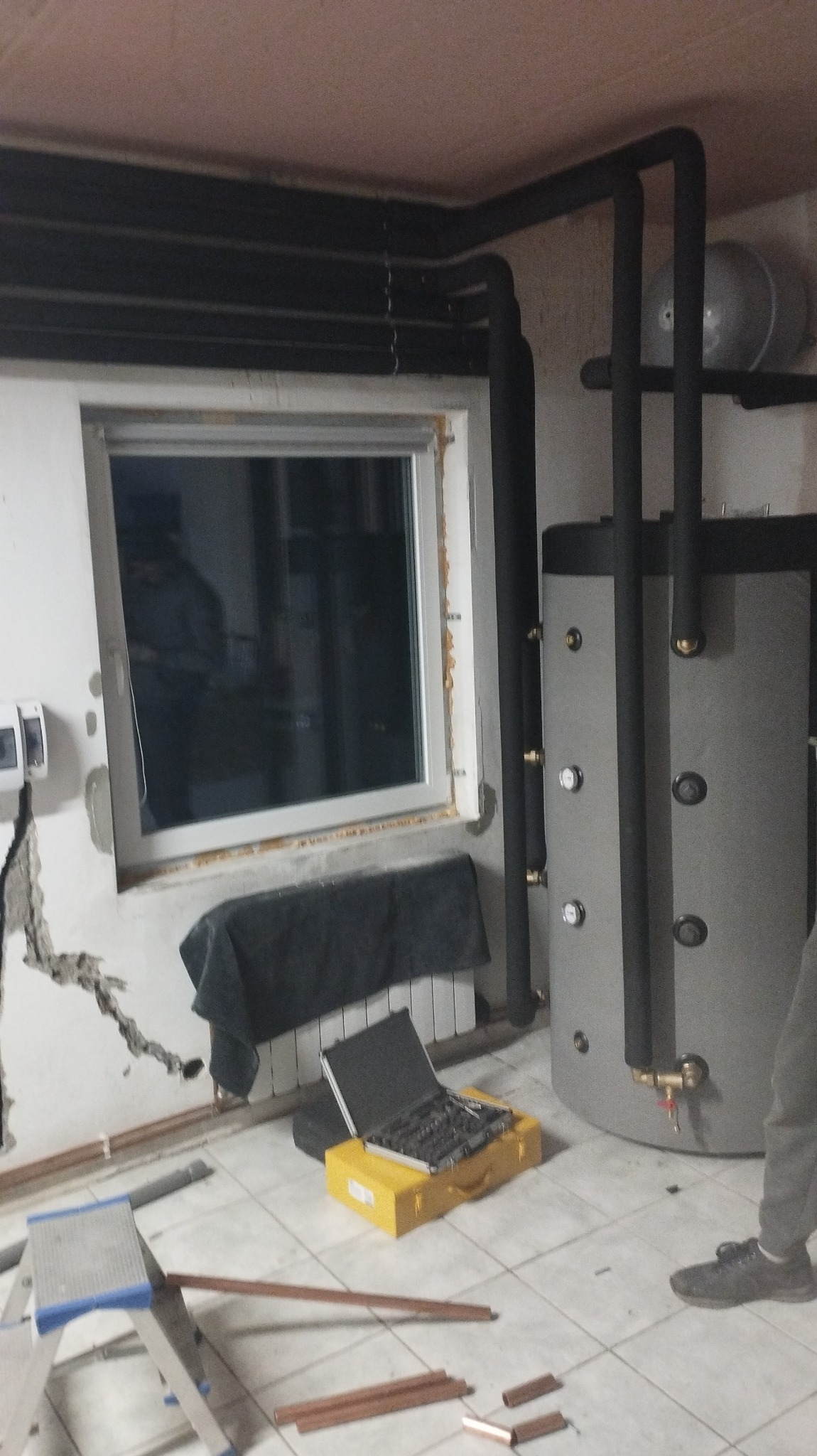 Szary zasobnik ciepłej wody użytkowej z czarnymi rurami instalacji grzewczej w trakcie montażu, widoczna drabina, walizka z narzędziami i miedziane rury.