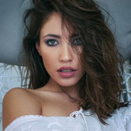 Tomasz Markieiwicz - Portret kobiety z zielonymi oczami, w białej bluzce z haftem, z mocnym makijażem oczu w odcieniach fioletu, leżącej w fotelu z szarymi poduszkami.