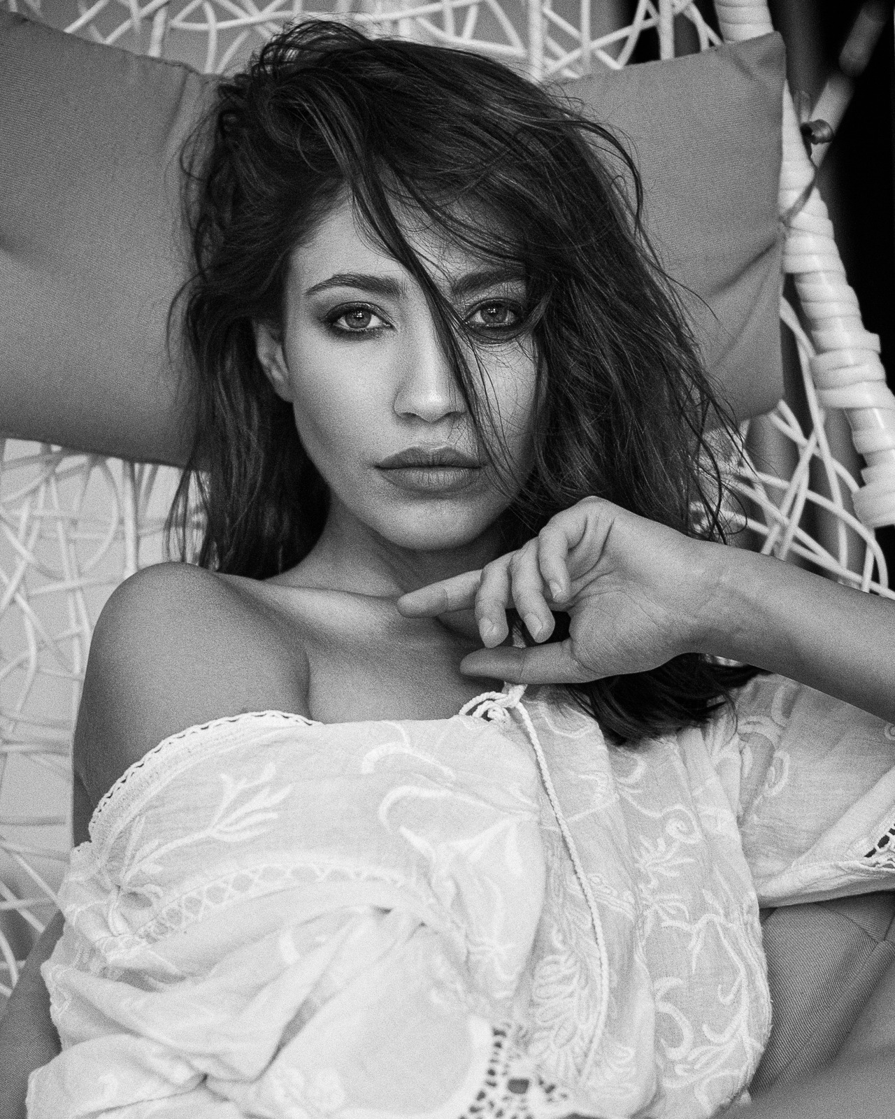 Czarno-biały portret kobiety w ażurowym fotelu, z delikatnym makijażem i bluzką z haftem, włosy opadają na twarz, dłoń przy ustach.