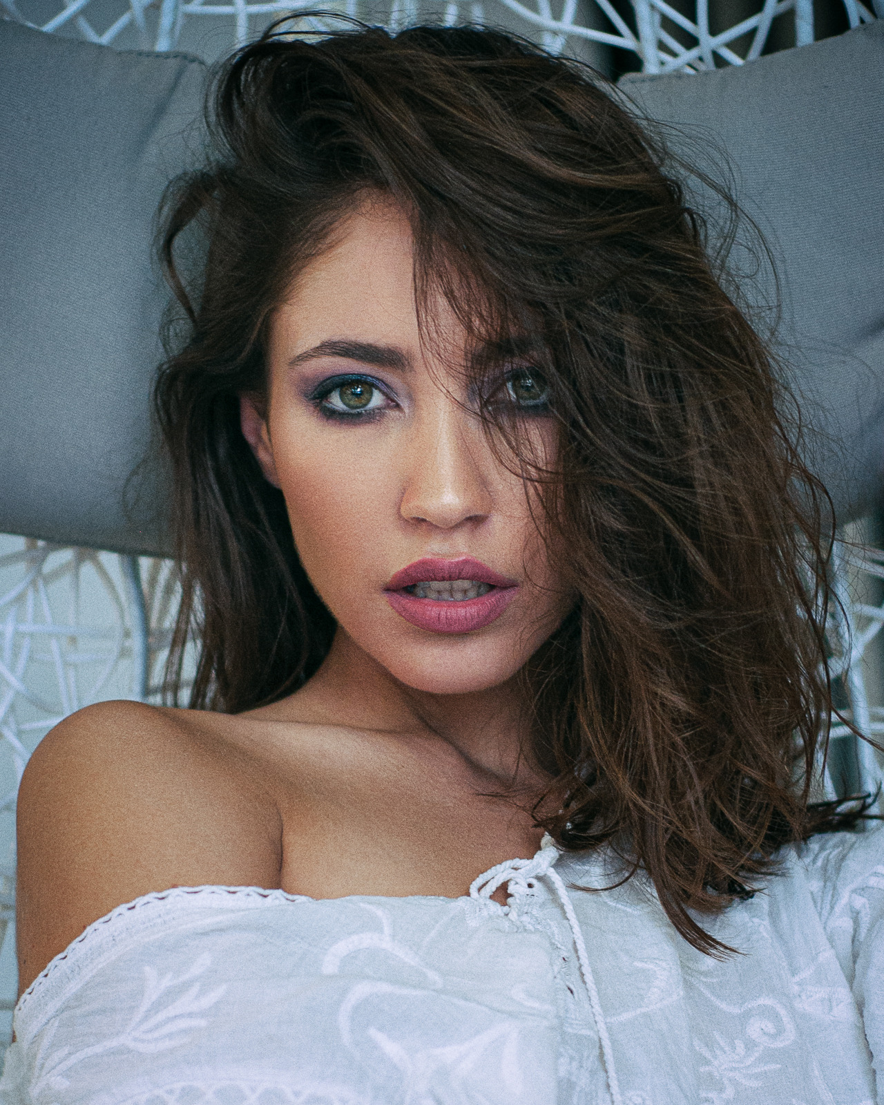 Portret kobiety z zielonymi oczami, w białej bluzce z haftem, z mocnym makijażem oczu w odcieniach fioletu, leżącej w fotelu z szarymi poduszkami.