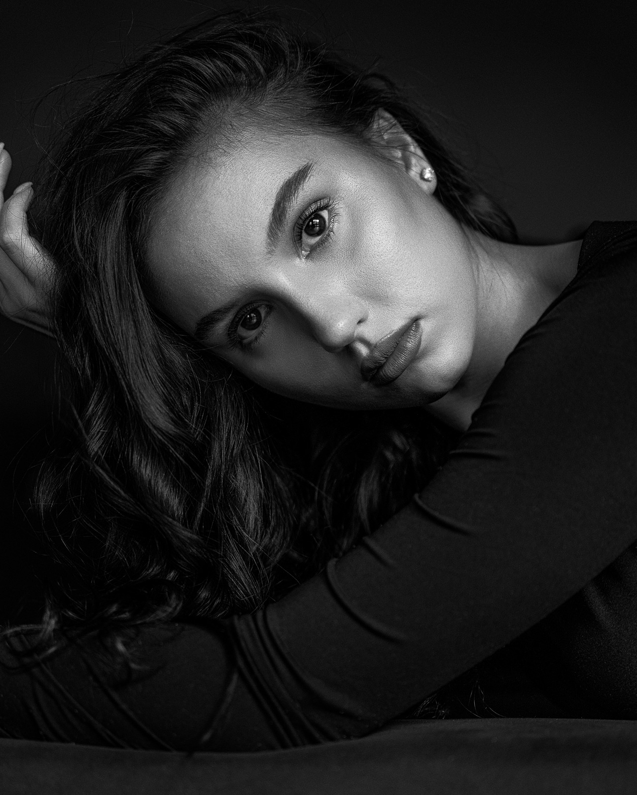 Czarno-biały portret kobiety z długimi, falującymi włosami, opierającej głowę na rękach, z delikatnym makijażem i kolczykiem w uchu, na ciemnym tle.