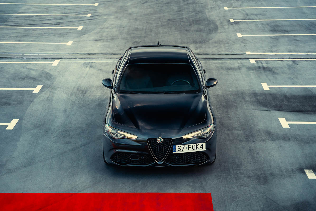 Czarna Alfa Romeo Giulia zaparkowana na szarym parkingu, widok z góry na przód samochodu, widoczna tablica rejestracyjna i czerwona linia na dole kadru.