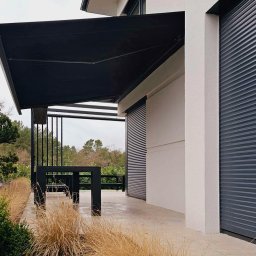 SPIN Bobko i Staniewski sp. j. - Czarna, prostokątna markiza zamontowana nad tarasem z widocznymi roletami zewnętrznymi na ścianie budynku w tle.