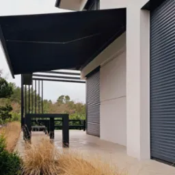 Czarna, prostokątna markiza zamontowana nad tarasem z widocznymi roletami zewnętrznymi na ścianie budynku w tle.