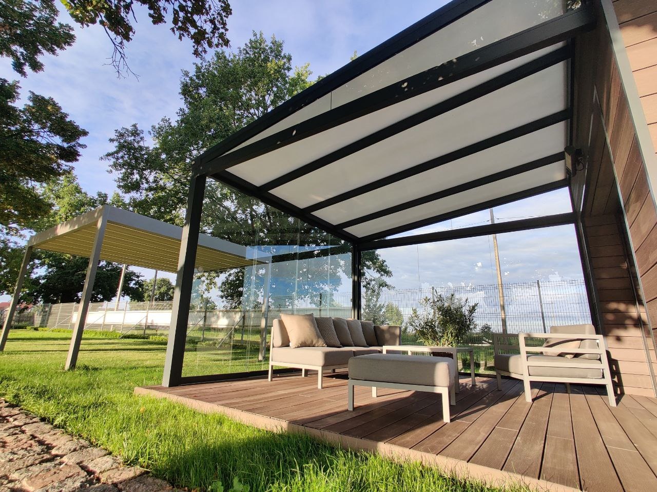 Nowoczesna konstrukcja zadaszenia tarasu z czarną ramą i białym dachem, wyposażona w szklane ściany boczne, stojąca na drewnianym podeście z meblami ogrodowymi w odcieniach szarości, w tle zielony...