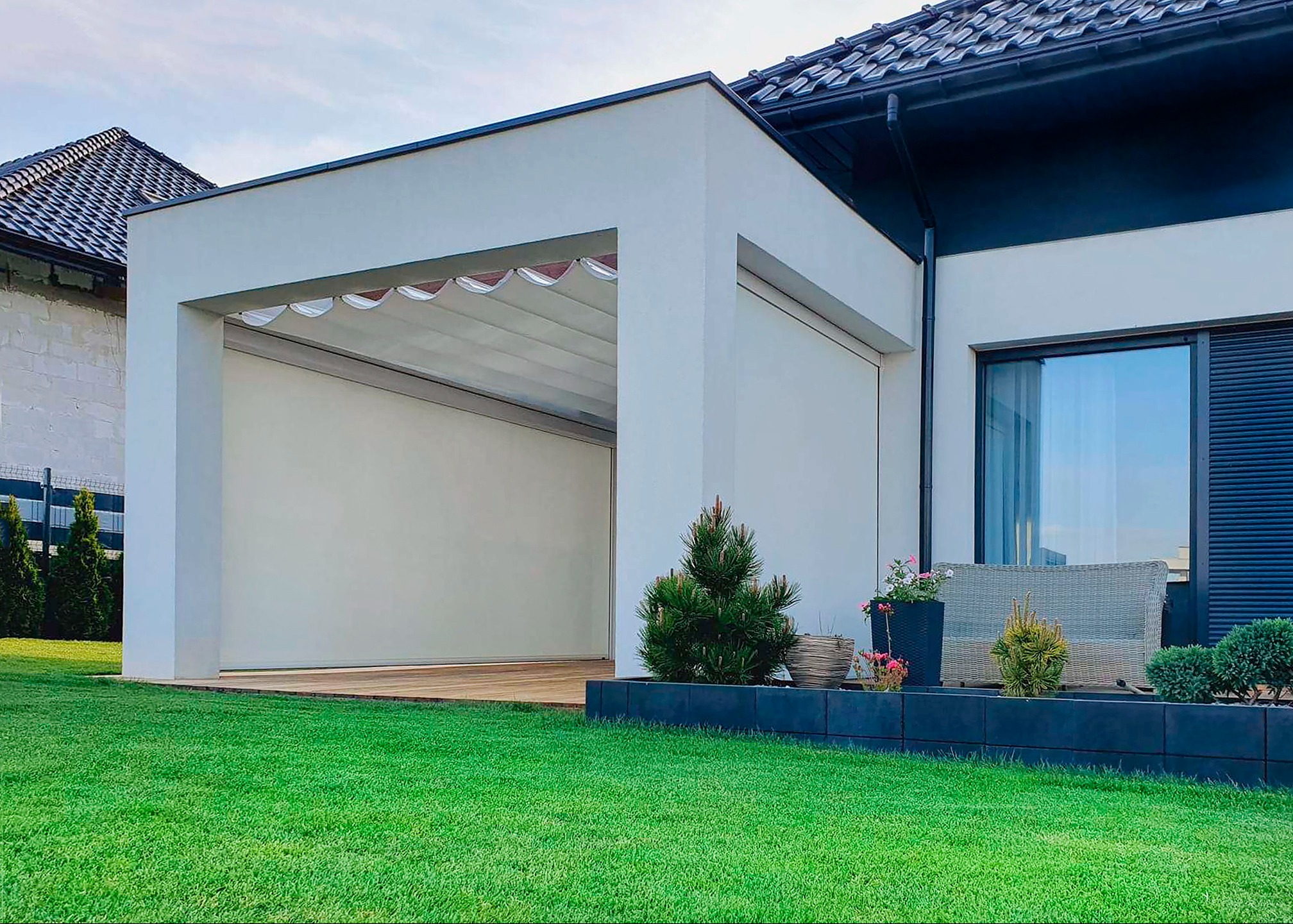 Nowoczesna pergola z zadaszeniem sterowanym ręcznie i osłonami bocznymi, wkomponowana w architekturę domu z zadbanym trawnikiem.