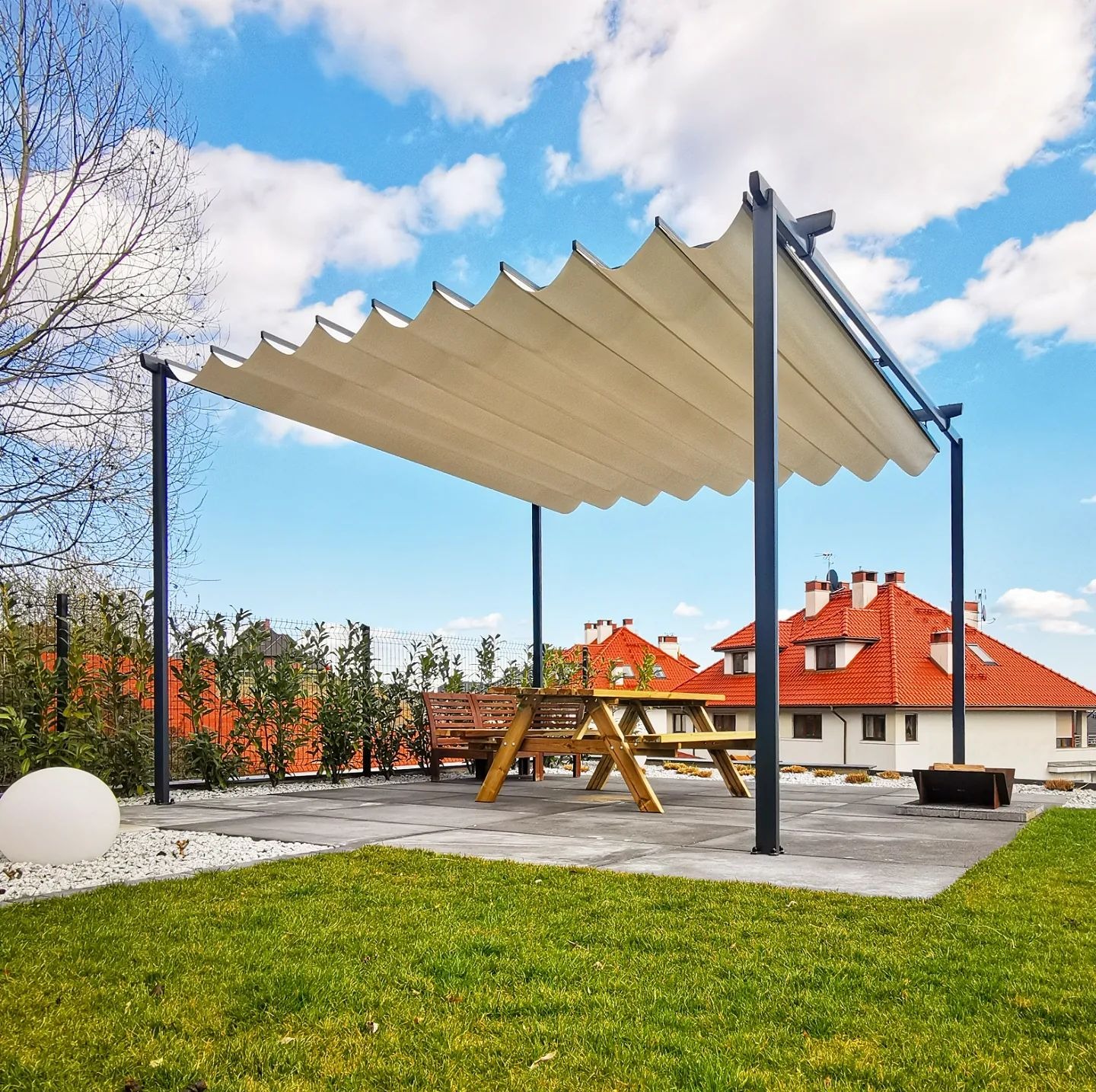 Nowoczesna konstrukcja z falistym, beżowym zadaszeniem na tarasie, chroniąca drewniany stół piknikowy; w tle trawnik, białe kamienie i dom z czerwoną dachówką na tle błękitnego nieba z chmurami.
