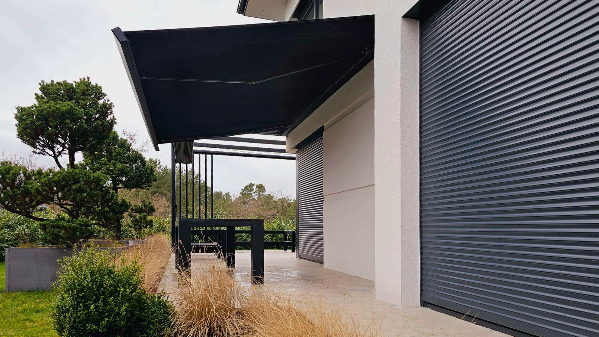 Czarna, prostokątna markiza zamontowana nad tarasem z widocznymi roletami zewnętrznymi na ścianie budynku w tle.
