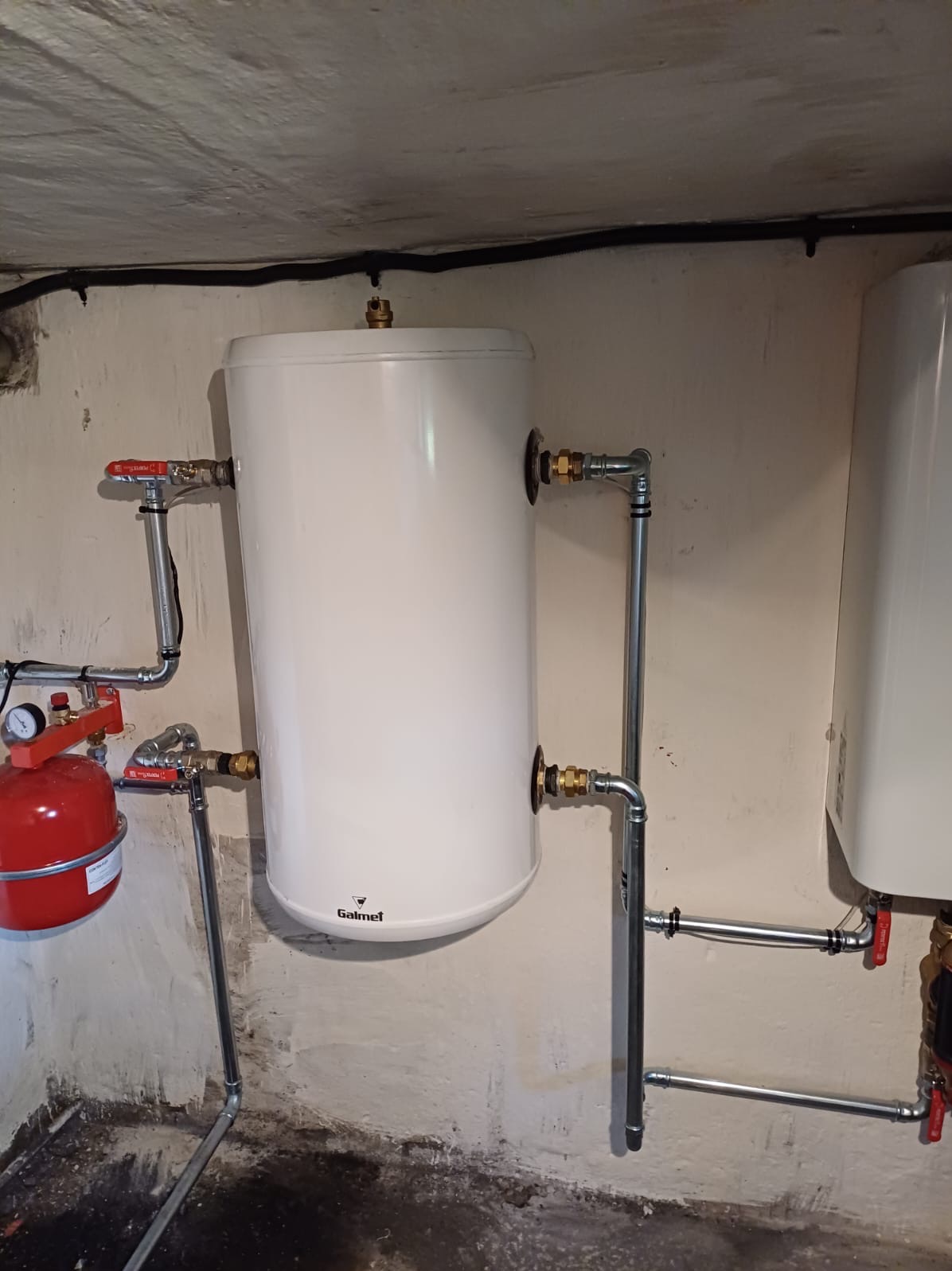 Montaż dwóch białych, cylindrycznych podgrzewaczy wody Galmet w piwnicy w Gorzowie Wielkopolskim, z widocznymi rurami i zaworami.