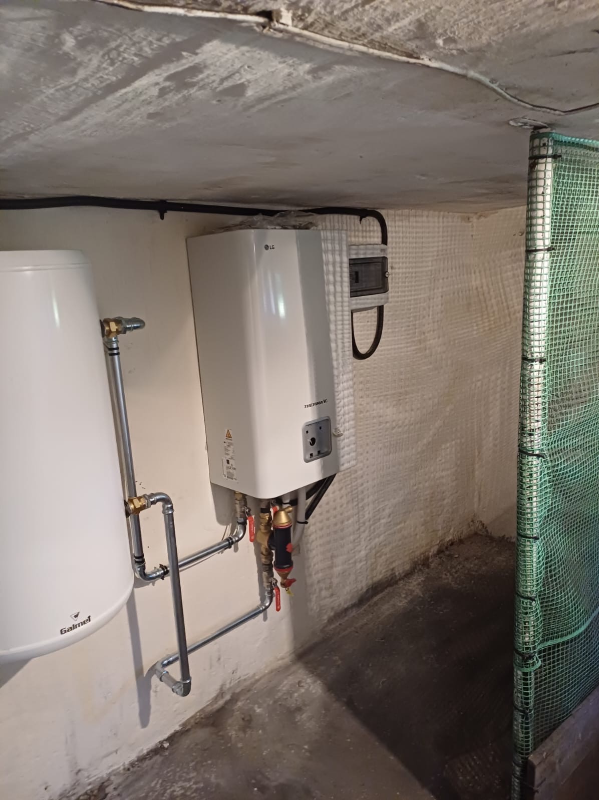 Biały zbiornik Galmet i pompa ciepła LG Therma V z podłączonymi rurami w surowym pomieszczeniu z betonowym sufitem i częściowo ocieplonymi ścianami.