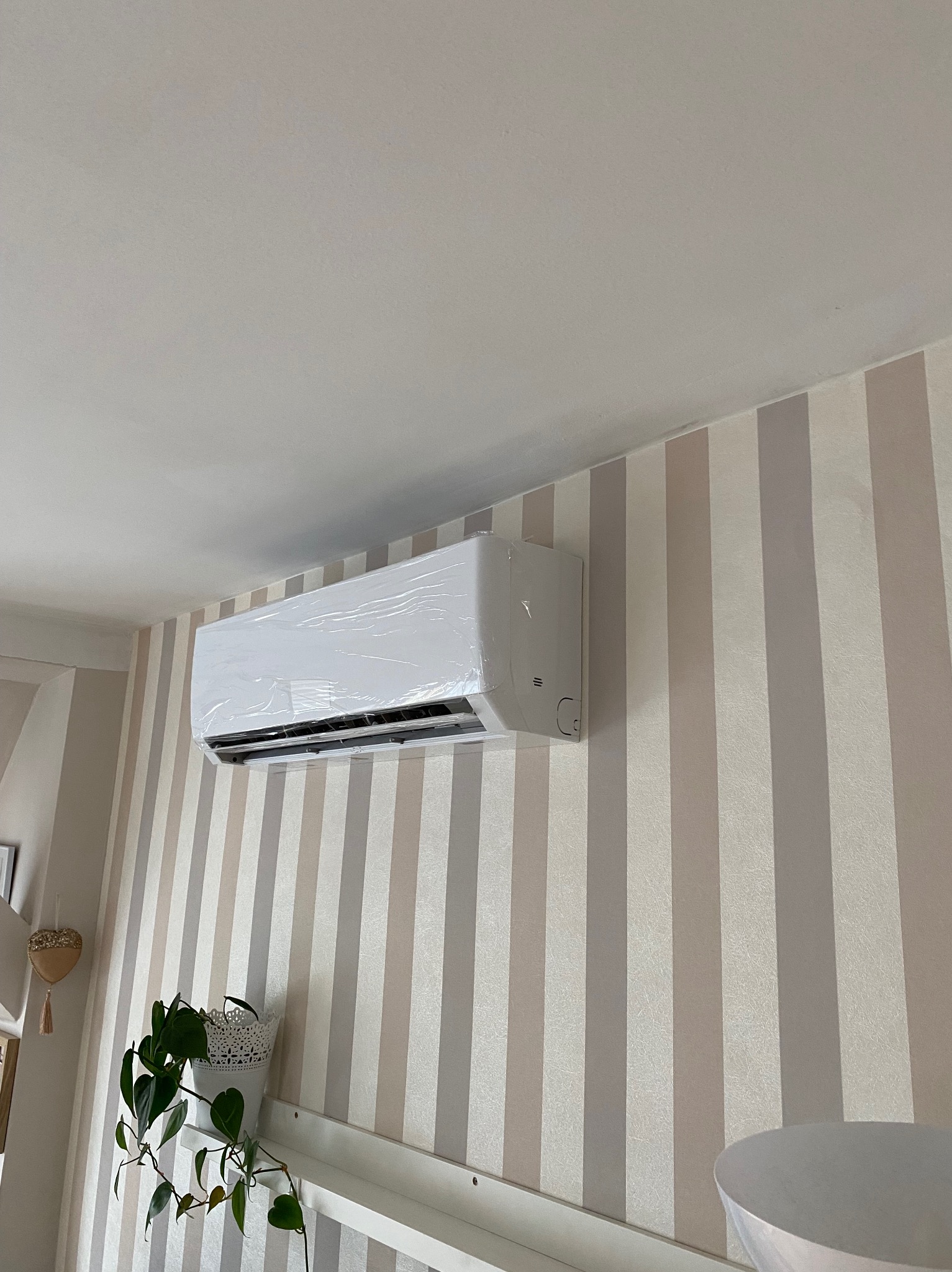 Nowy, biały klimatyzator ścienny, jeszcze w folii ochronnej, zamontowany w pokoju z tapetą w pionowe pasy.