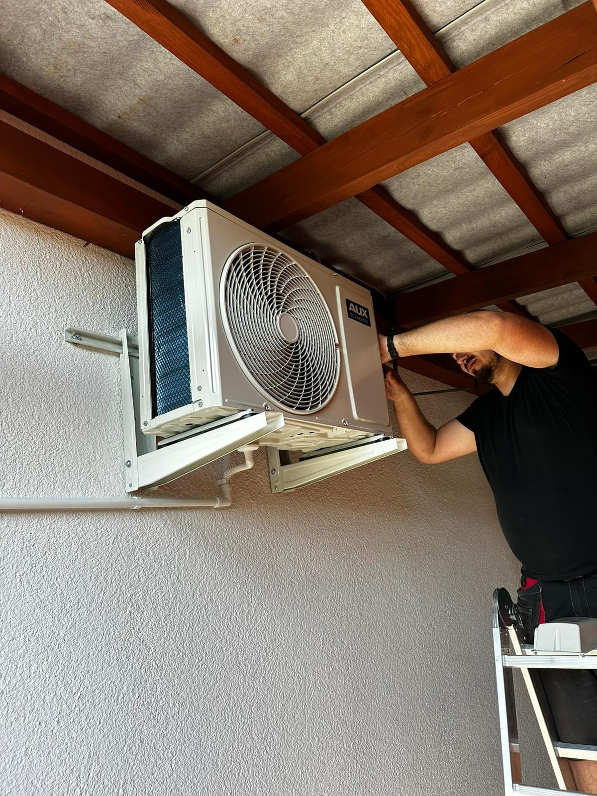 Montaż zewnętrznej jednostki klimatyzacji marki AUX na wspornikach, widoczny instalator na drabinie, ściana o fakturze baranka i drewniany strop.