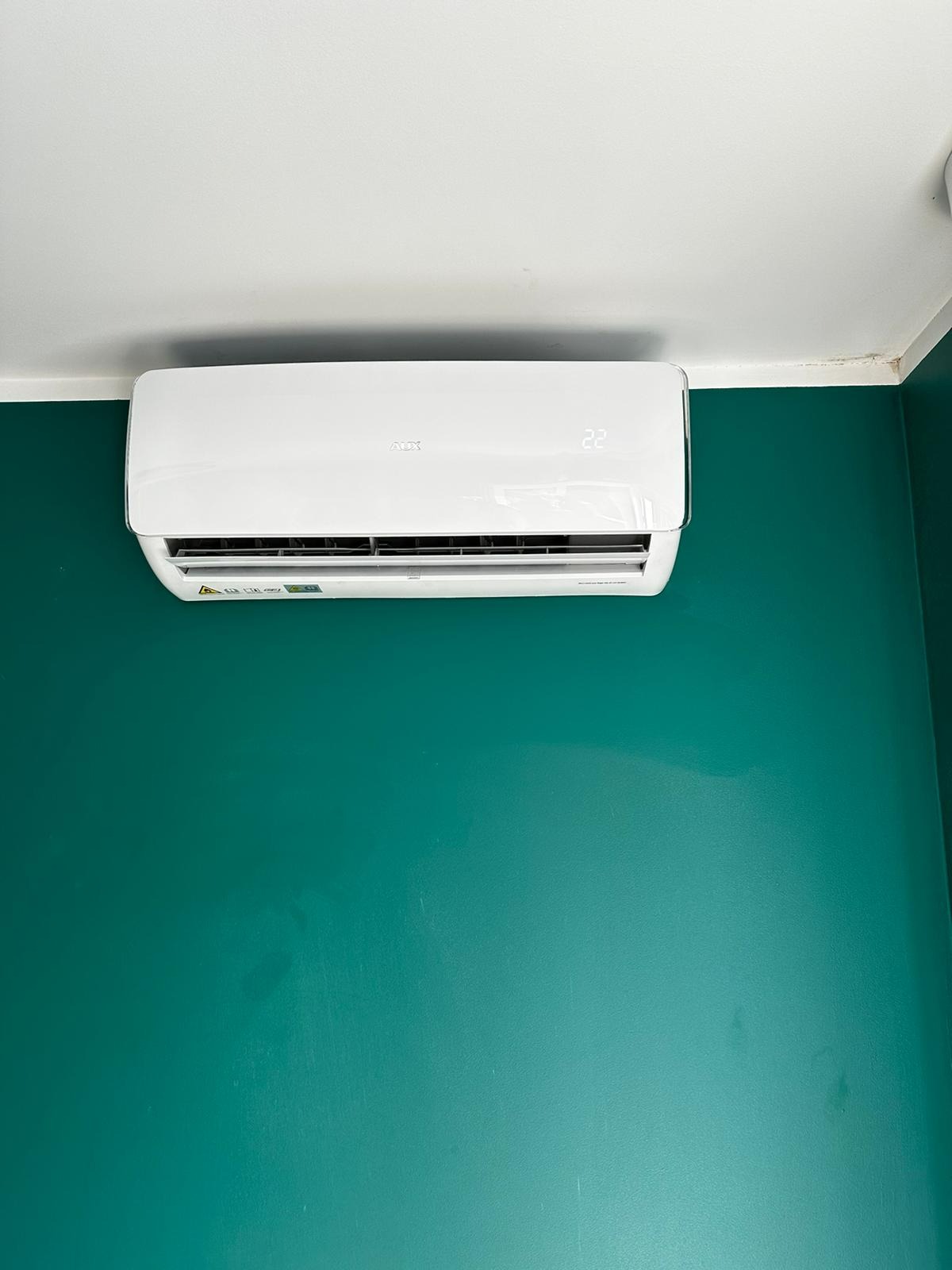 Biała klimatyzacja ścienna marki AUX w minimalistycznym wnętrzu z zieloną ścianą, wyświetlająca temperaturę 22 stopnie.