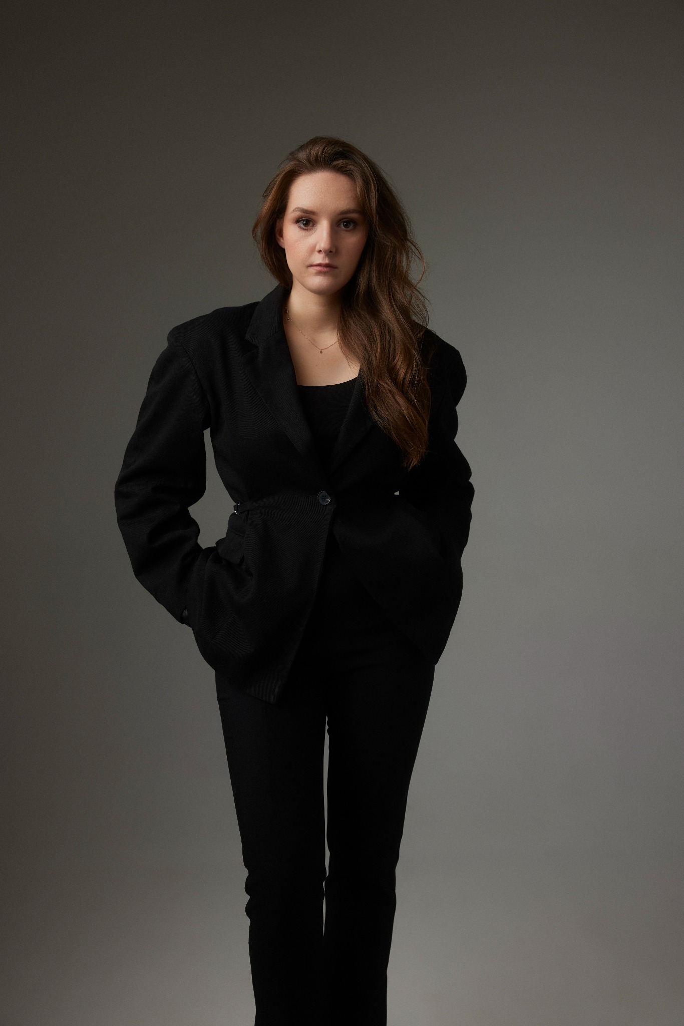 Portret kobiety w czarnym garniturze na szarym tle, z rękami w kieszeniach, patrzącej wprost w obiektyw.