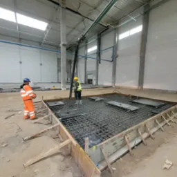 Dwóch pracowników w hali przemysłowej przygotowuje szalunek i zbrojenie pod wylewkę betonową; widoczna pompa do betonu.