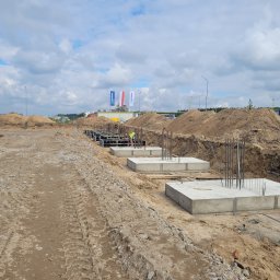 Kamil Malcher - Przygotowane betonowe stopy fundamentowe z wystającymi prętami zbrojeniowymi na placu budowy, widoczny robotnik w kamizelce odblaskowej, w tle maszt z flagami.