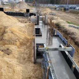 Wykop z szalunkiem pod fundament z zalanym betonem, wystające pręty zbrojeniowe, piasek i budynki mieszkalne w tle.