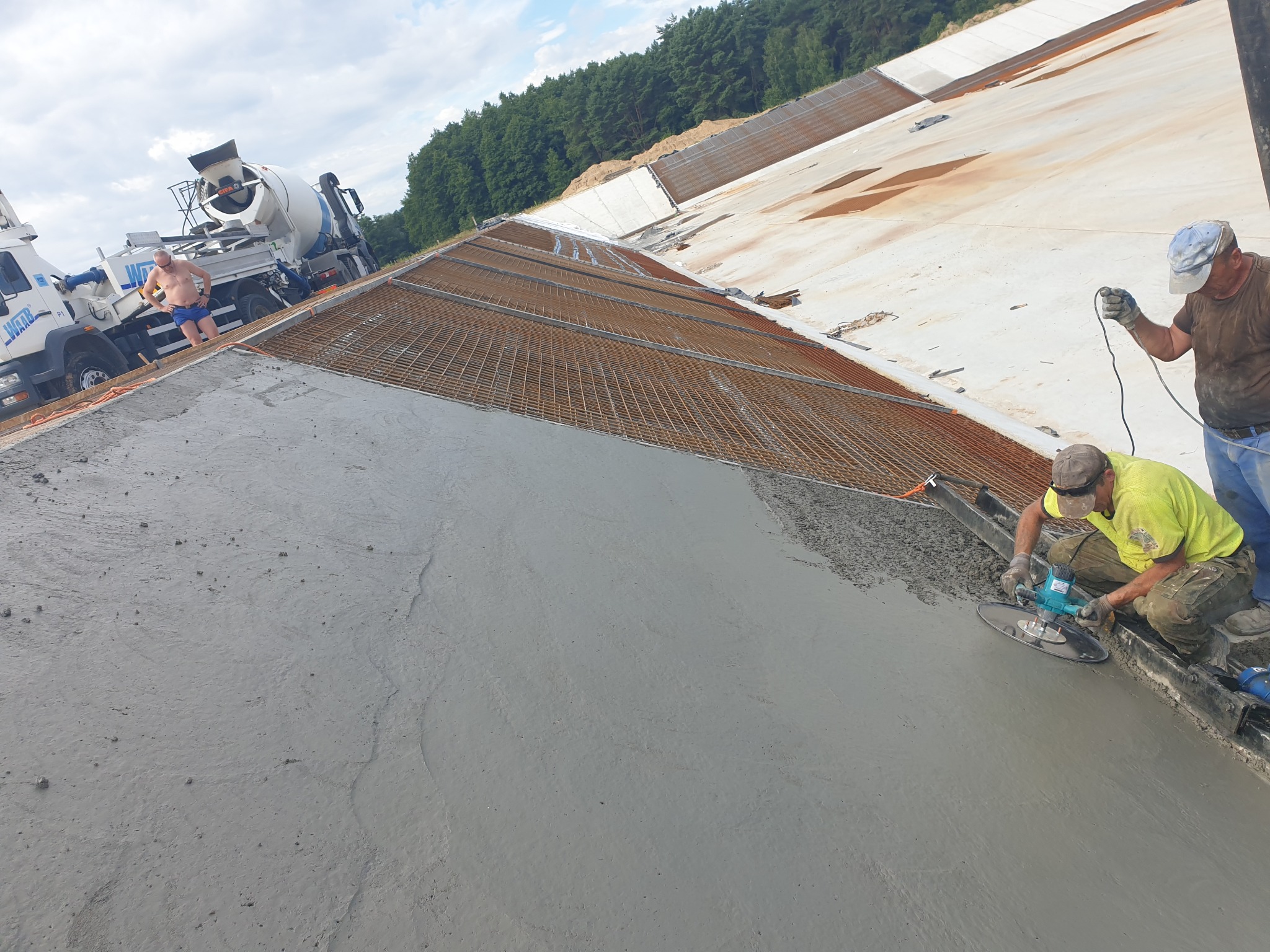 Wygładzanie świeżo wylanej betonowej powierzchni zbrojonej siatką, w tle betoniarki i pracownik bez koszulki.