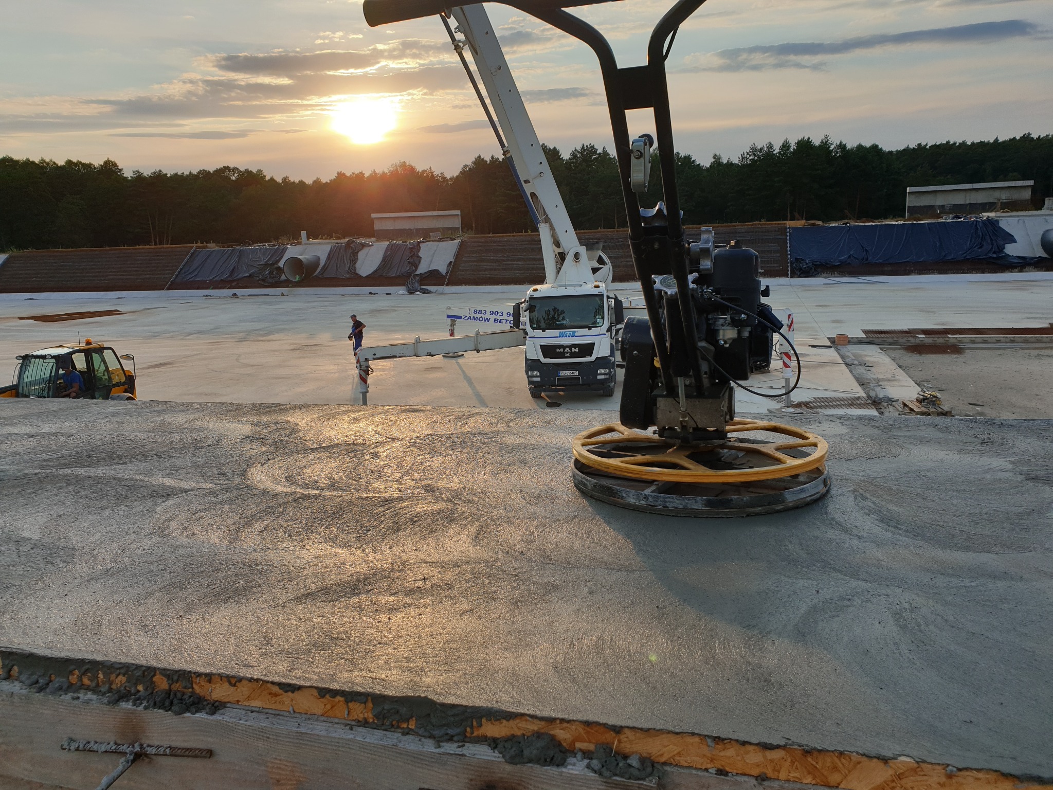 Świeżo wylana, gładzona mechanicznie posadzka betonowa w promieniach zachodzącego słońca, z widoczną maszyną do zacierania betonu, pompą do betonu i operatorem w tle.