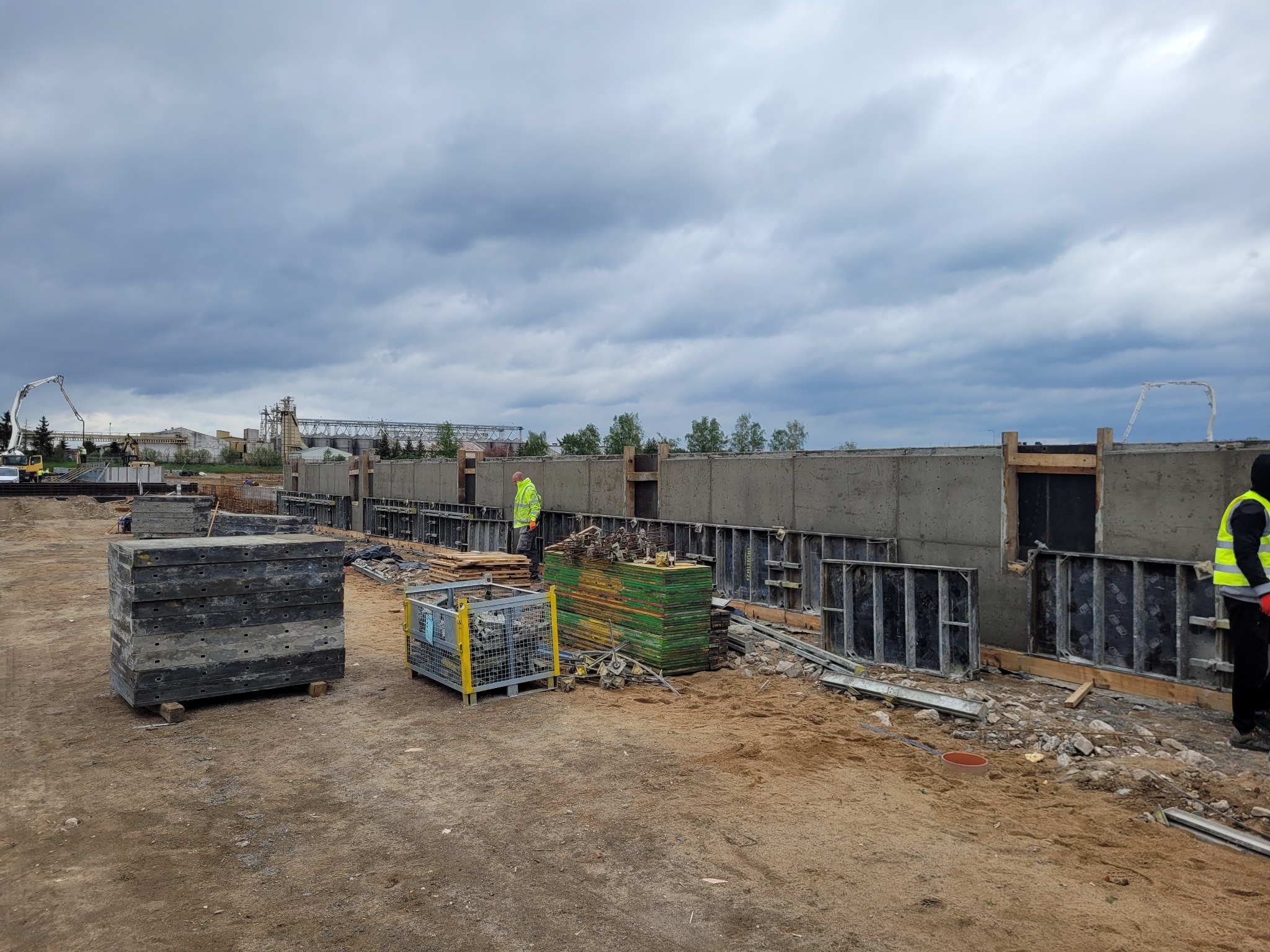 Szare betonowe ściany szalunkowe w trakcie budowy, widoczny pracownik w żółtej kamizelce, materiały budowlane i sprzęt do betonu w tle pod pochmurnym niebem.