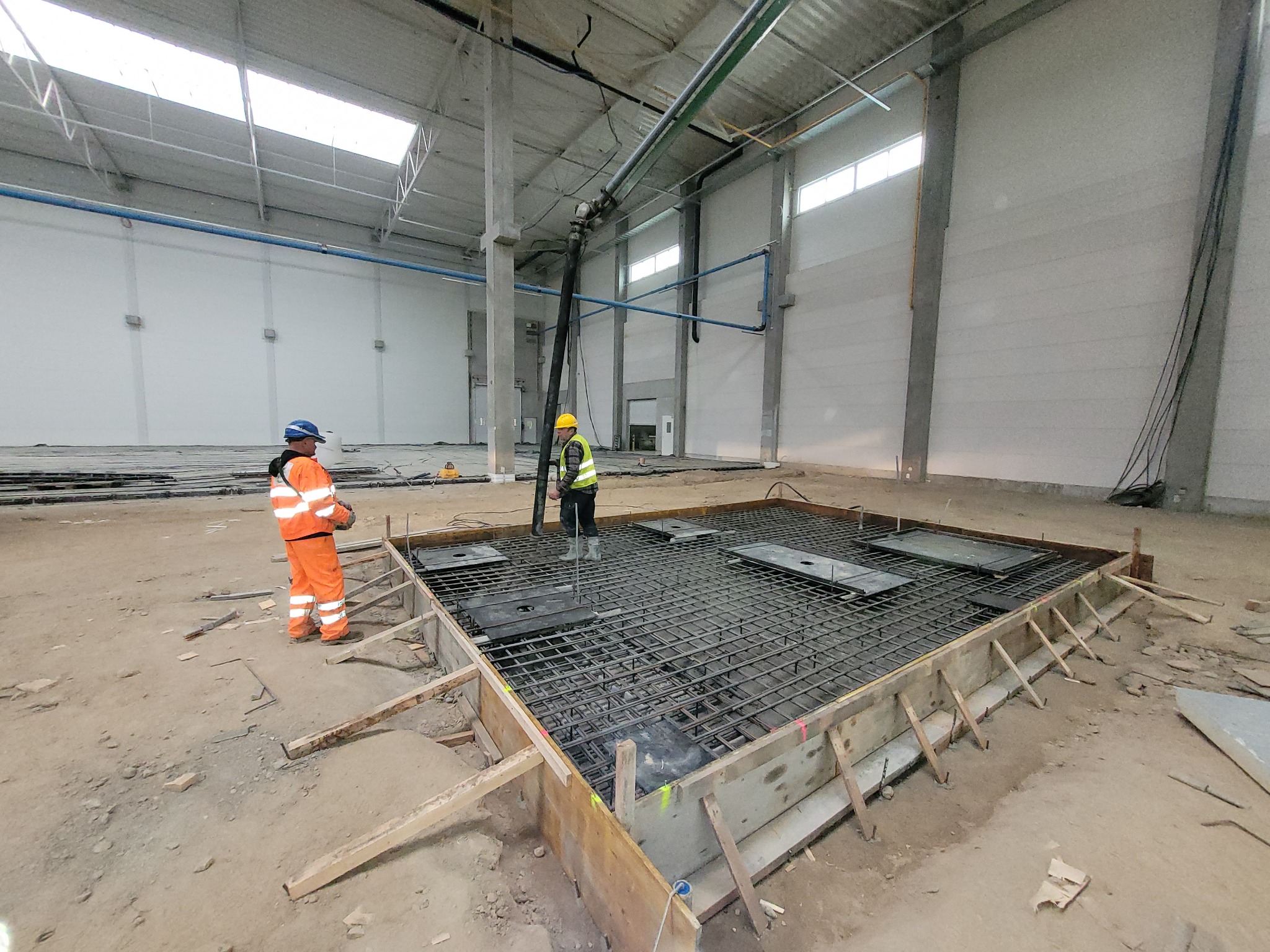 Dwóch pracowników w hali przemysłowej przygotowuje szalunek i zbrojenie pod wylewkę betonową; widoczna pompa do betonu.