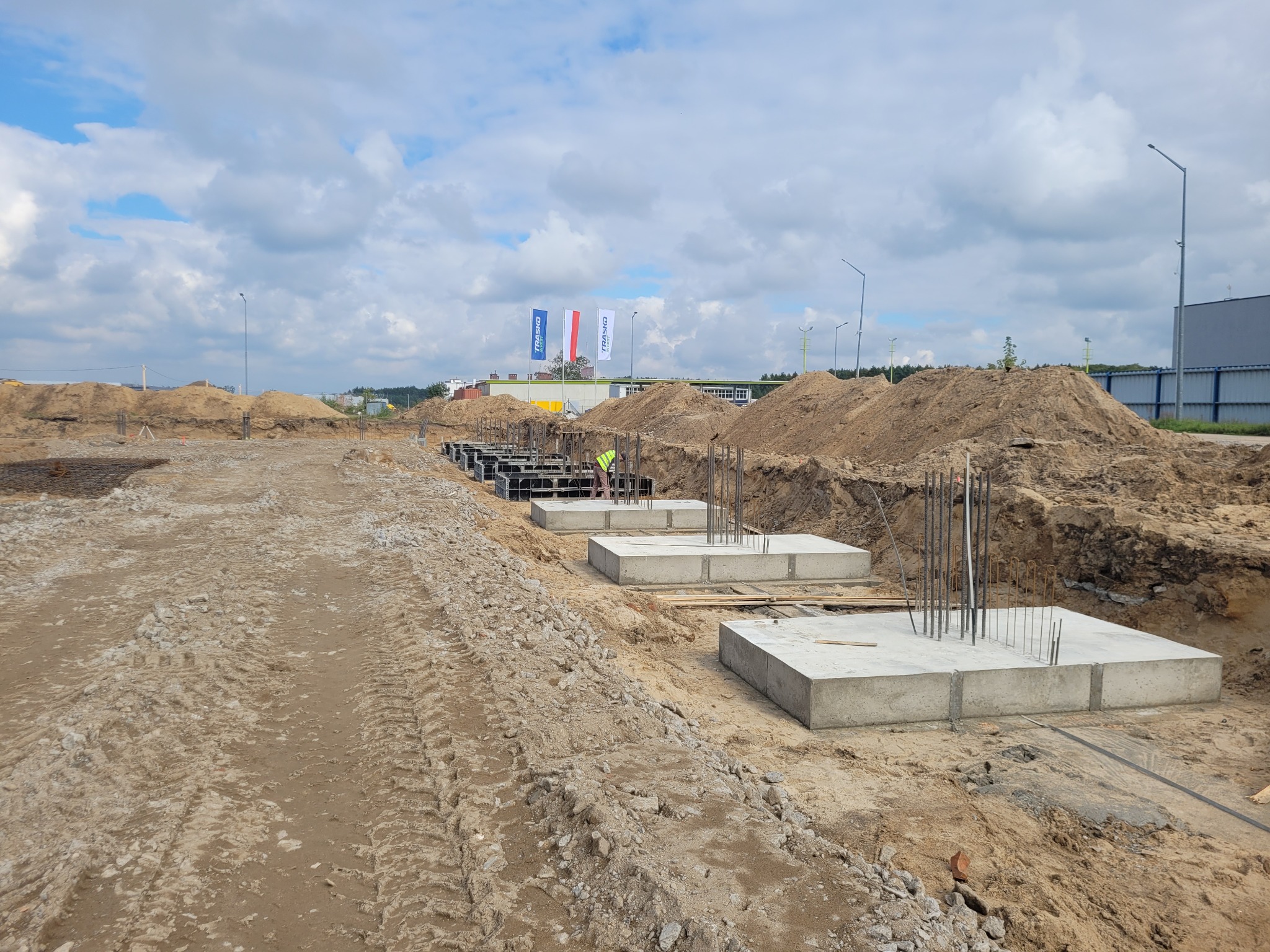 Przygotowane betonowe stopy fundamentowe z wystającymi prętami zbrojeniowymi na placu budowy, widoczny robotnik w kamizelce odblaskowej, w tle maszt z flagami.