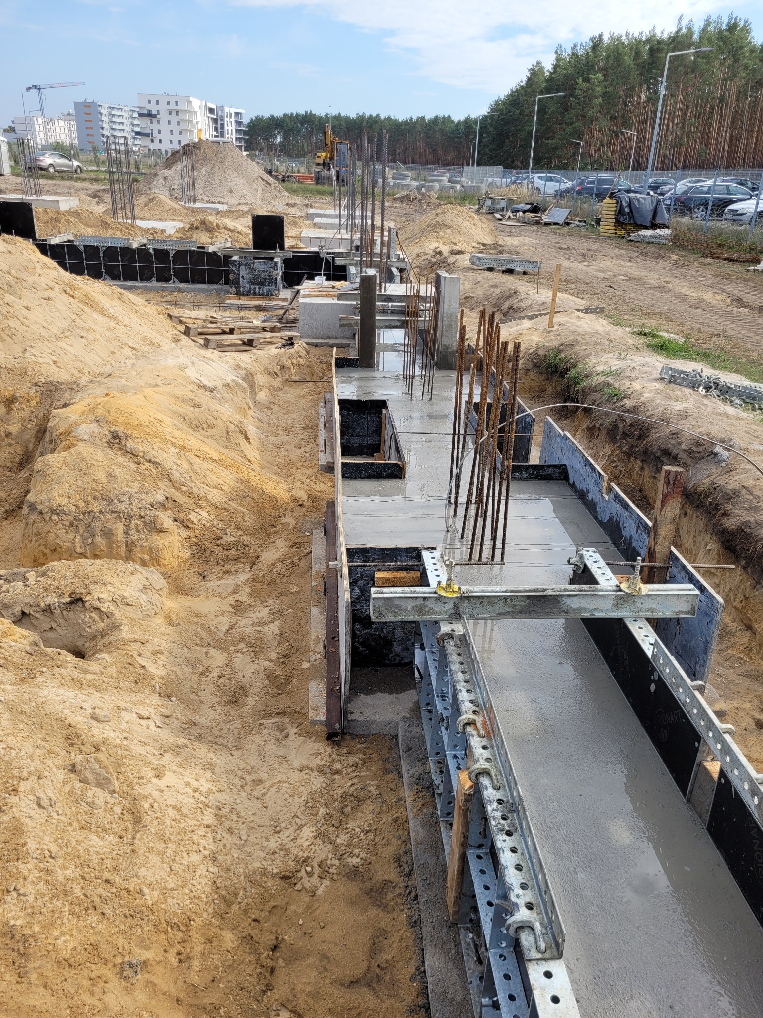 Wykop z szalunkiem pod fundament z zalanym betonem, wystające pręty zbrojeniowe, piasek i budynki mieszkalne w tle.