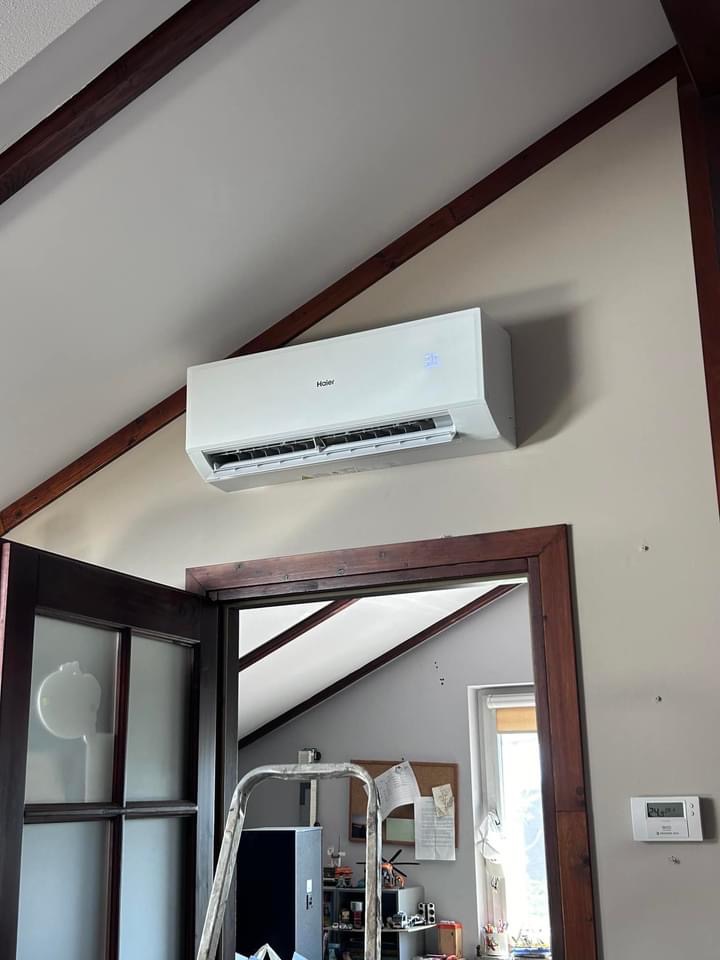 Biały klimatyzator Haier zamontowany pod skośnym sufitem w domu, widoczna drabina i drzwi w tle.