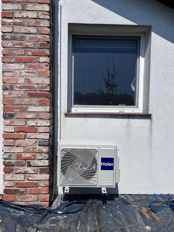 Zewnętrzna jednostka klimatyzacji Haier zamontowana na elewacji budynku obok okna i ceglanego narożnika, widoczne folie ochronne na dachu.