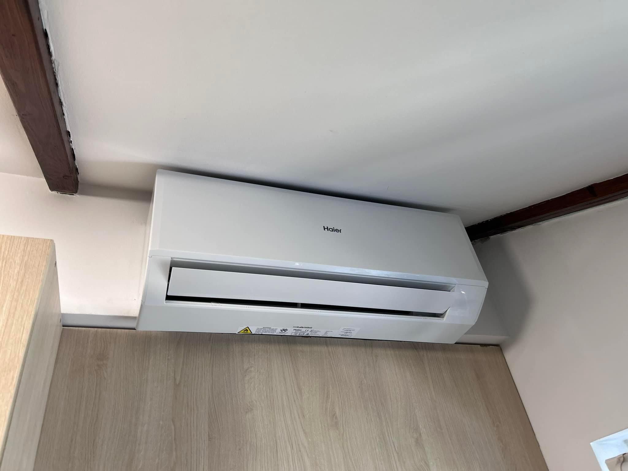 Biała klimatyzacja Haier zamontowana przy suficie z widoczną drewnianą belką, zainstalowana w pomieszczeniu z panelami drewnopodobnymi, widok od dołu.