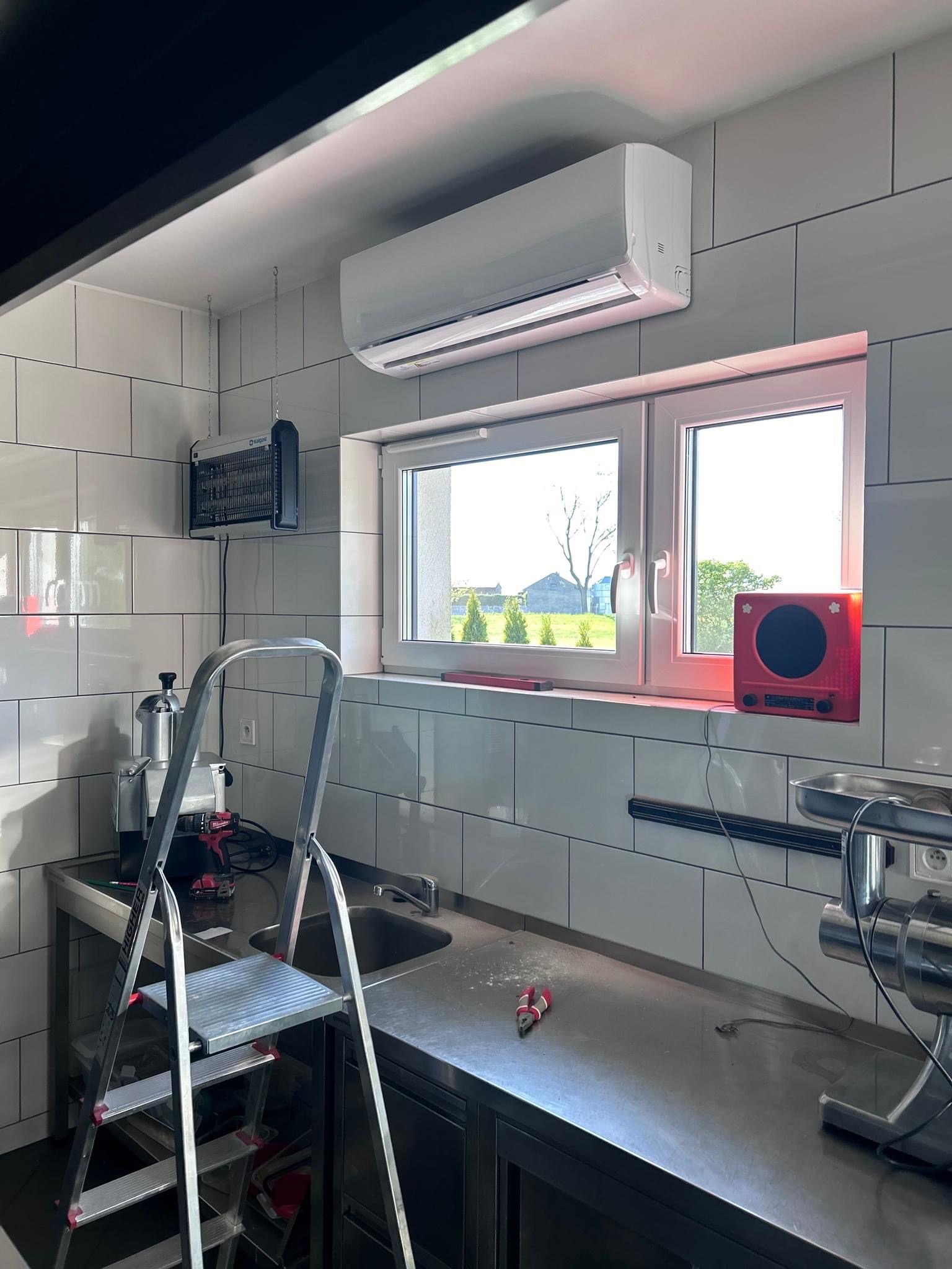 Wnętrze kuchni z zamontowanym białym klimatyzatorem ściennym, drabina aluminiowa, narzędzia i sprzęt kuchenny na metalowym blacie.