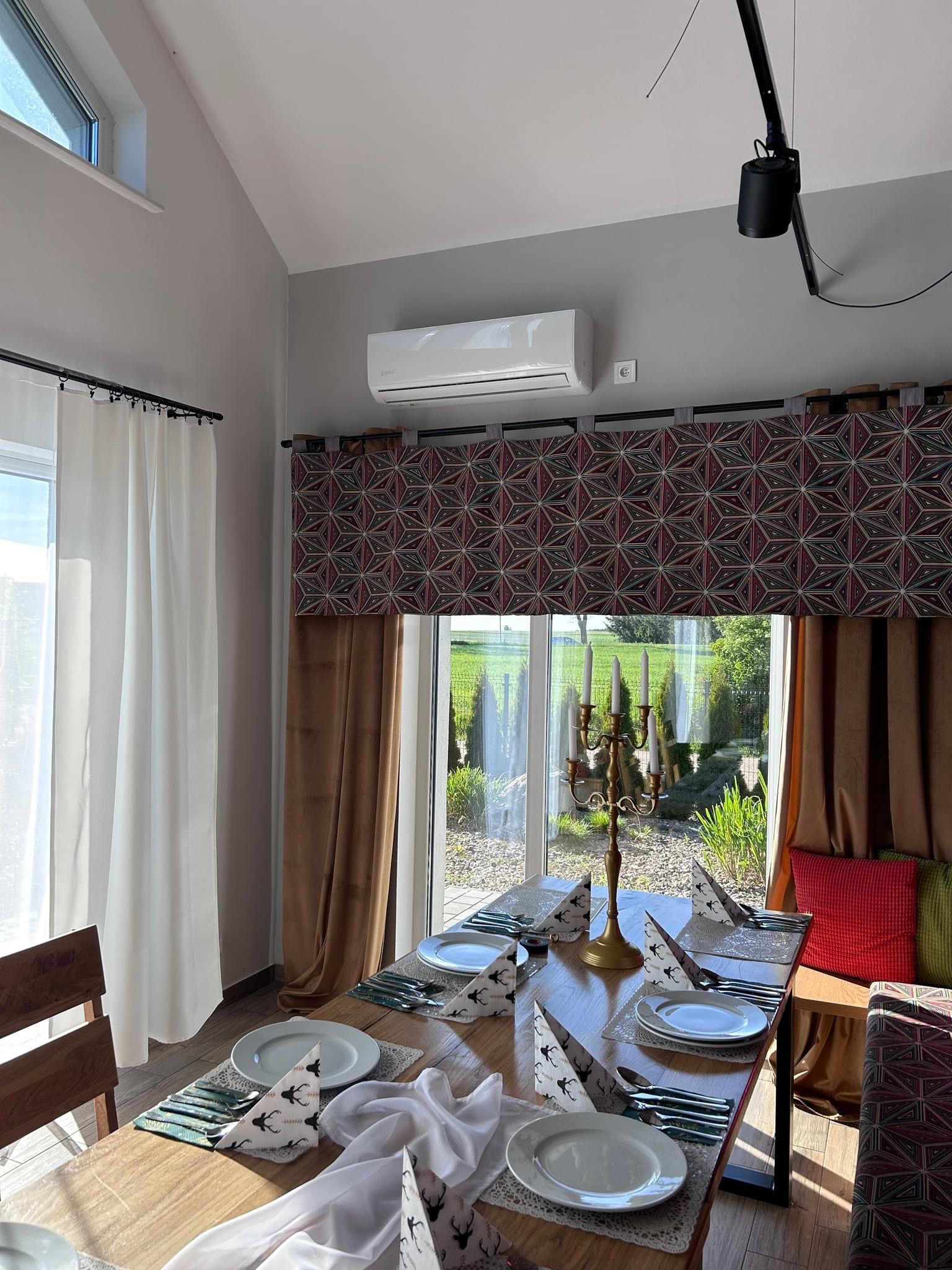 Wnętrze jadalni z zainstalowanym klimatyzatorem ściennym, widocznym nad oknem z dekoracyjną zasłoną i elegancko nakrytym stołem. Dyskretna, biała jednostka klimatyzacyjna wkomponowana w wystrój...