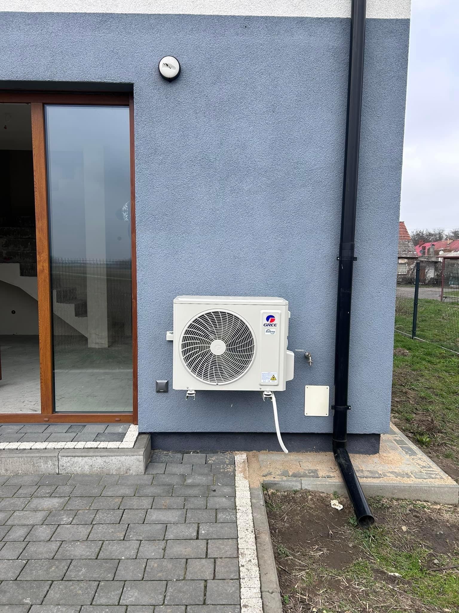 Zewnętrzna jednostka klimatyzacji marki Gree zamontowana na ścianie domu z szarą elewacją, widoczna rynna spustowa i fragment kostki brukowej.