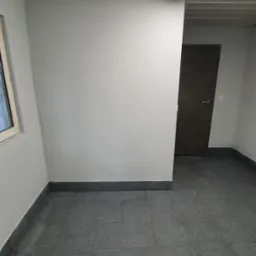 Pomalowane na szaro wnętrze pomieszczenia z szarą podłogą z płytek, widoczne okno i drzwi, sufit z widocznymi elementami konstrukcyjnymi i lampą jarzeniową.