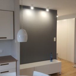 Rafdi Usługi Remontowe Rafał Ditner - Szara ściana z oświetleniem punktowym w nowoczesnym salonie, biały stół i krzesła na pierwszym planie, butelka wody na białej komodzie, drewniana podłoga.