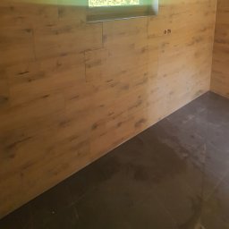 Rafdi Usługi Remontowe Rafał Ditner - Ściana wyłożona panelami imitującymi drewno, z widocznym gniazdkiem elektrycznym i małym oknem, połączona z ciemną, błyszczącą podłogą z płytek.