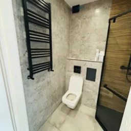 Wykończona łazienka z płytkami imitującymi kamień, czarnym grzejnikiem drabinkowym, toaletą wiszącą i kabiną prysznicową z czarnymi profilami i drewnopodobnymi panelami.