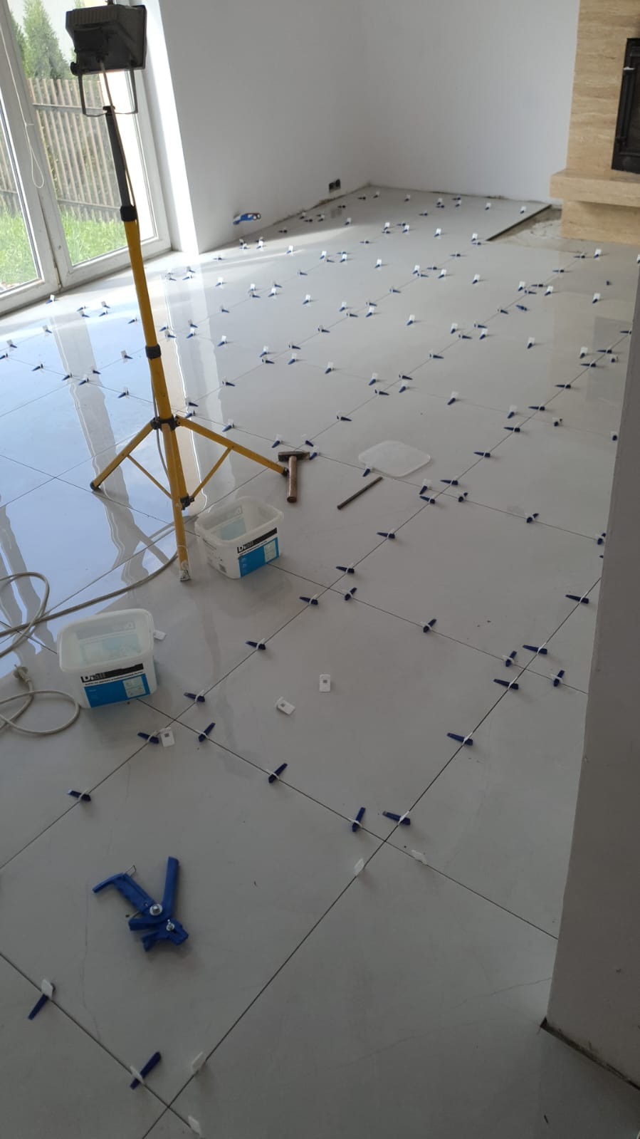 Układanie jasnych, kwadratowych płytek podłogowych z użyciem krzyżyków dystansowych, widoczna lampa budowlana, pojemniki z klejem, młotek i narzędzie do dociskania płytek.