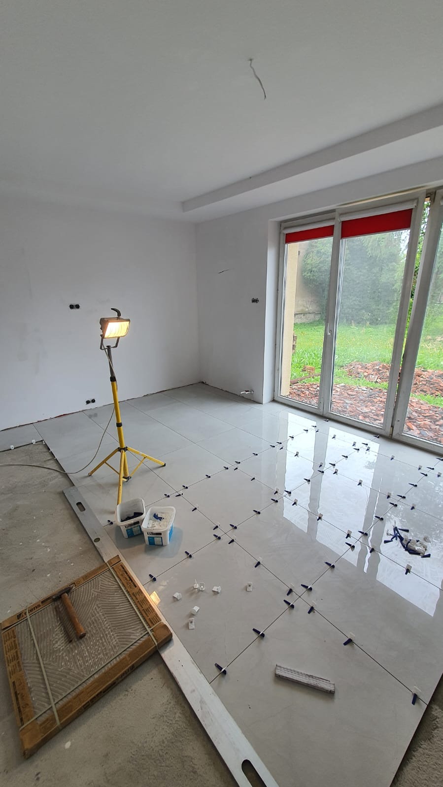 Proces układania jasnych, błyszczących płytek podłogowych w przestronnym, jasnym pomieszczeniu z dużym oknem; widoczna lampa robocza na statywie, poziomica, pojemniki z klejem i krzyżyki dystansowe.