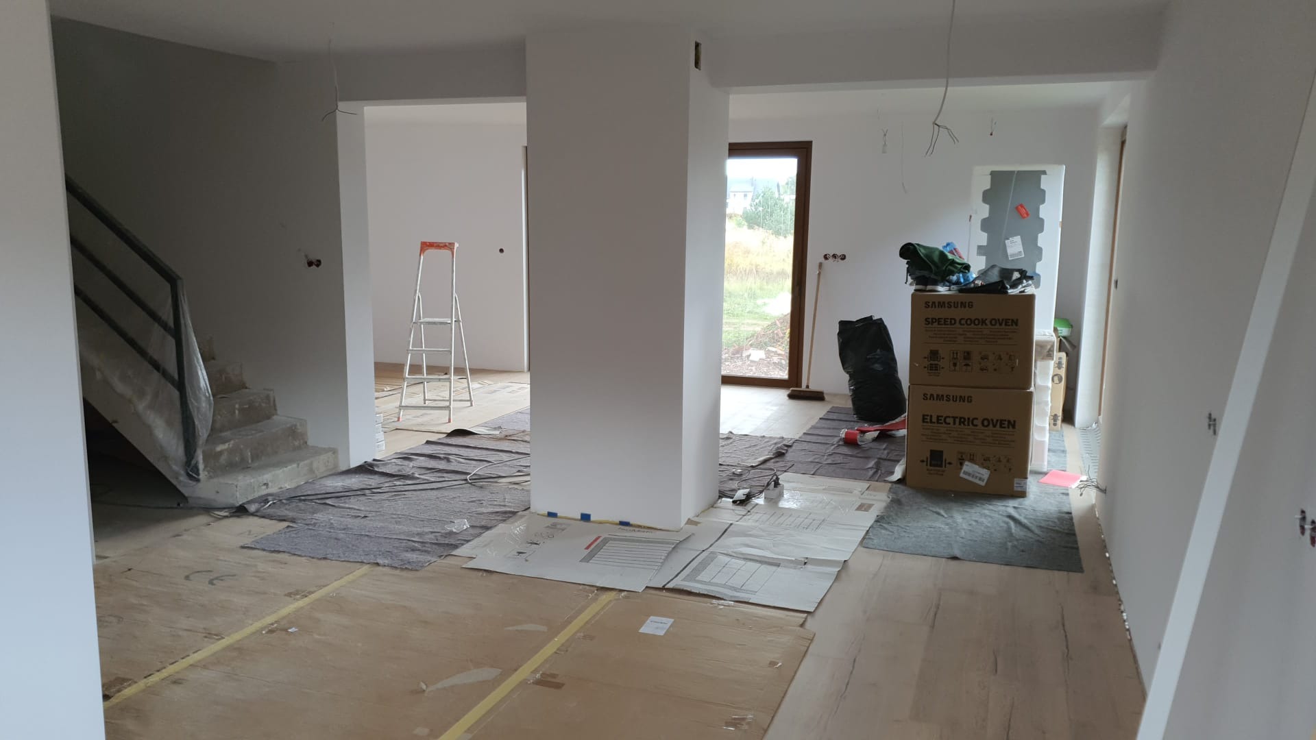 Wnętrze w trakcie prac wykończeniowych: surowe schody zabezpieczone folią, podłoga częściowo zakryta kartonem i folią ochronną, stojące kartony z piekarnikami Samsung, widoczna drabina i białe...