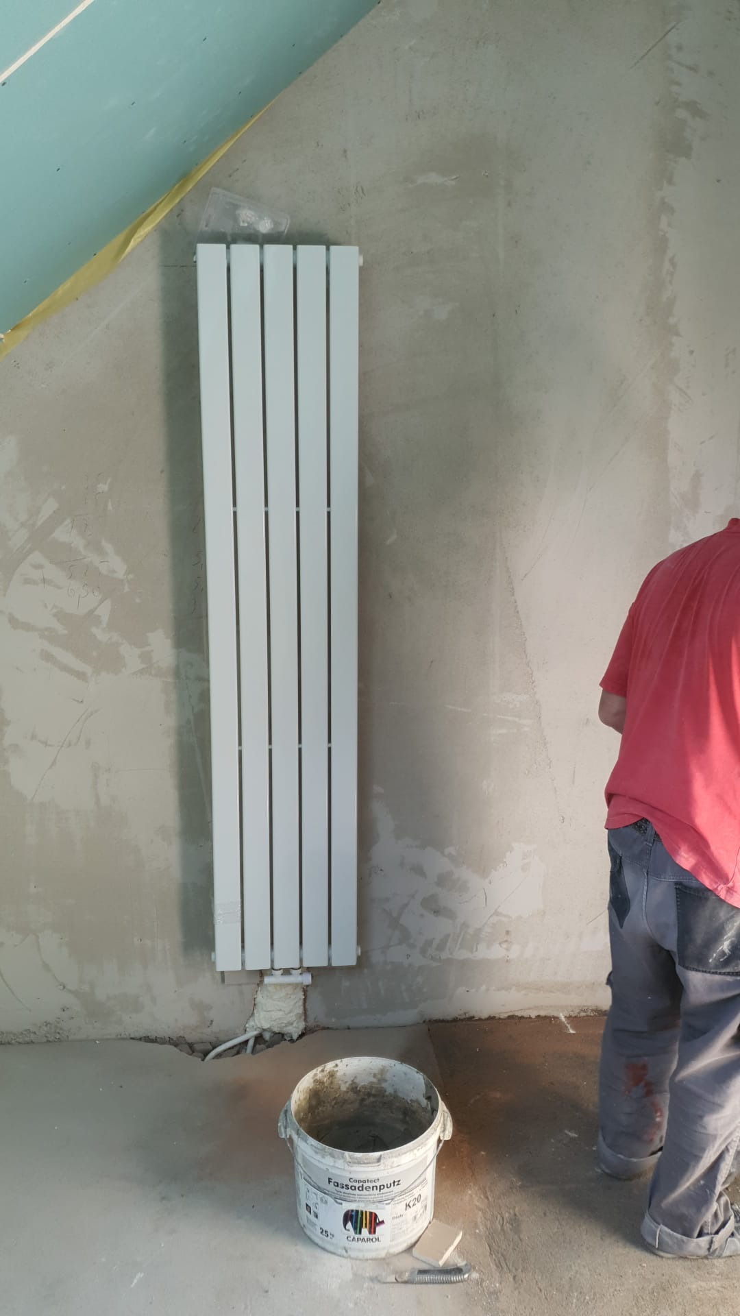 Montaż grzejnika panelowego na surowej ścianie w trakcie remontu, widoczny kubeł z zaprawą fasadową Caparol, ściany przygotowane pod gładzie, fragment sufitu z płyt gipsowo-kartonowych.