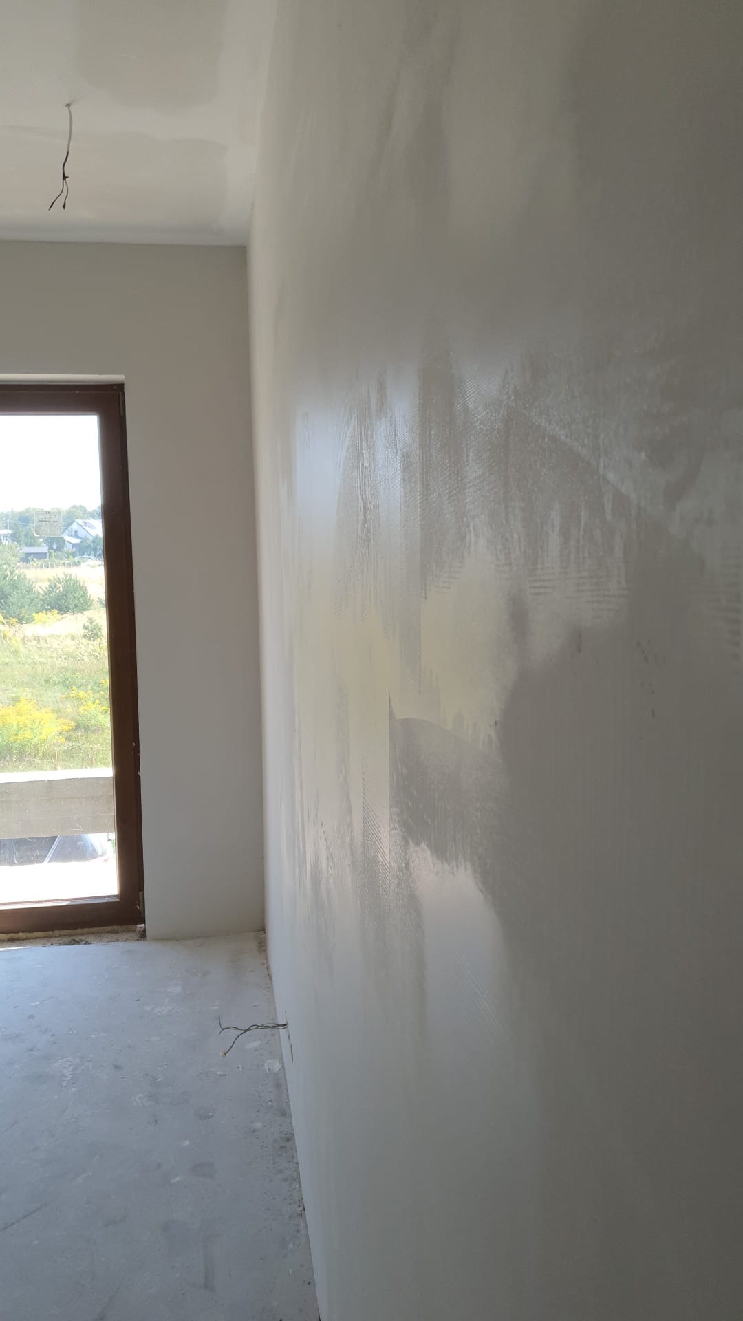 Świeżo położona gładź szpachlowa na ścianie w trakcie schnięcia, widoczne ślady po szpachli, surowy stan, brak listew przypodłogowych, wystające przewody elektryczne.