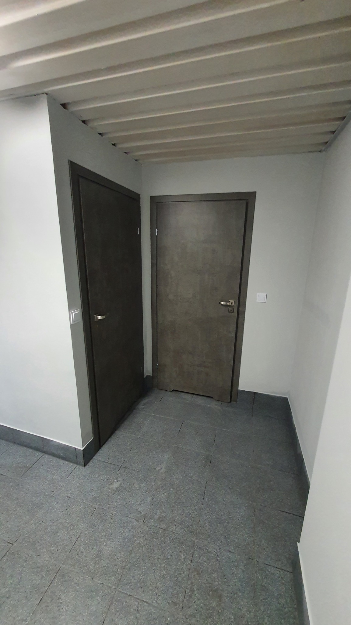 Dwa ciemnoszare drzwi wewnętrzne w rogu korytarza z szarą podłogą z płytek i białymi ścianami, widoczne listwy przypodłogowe.