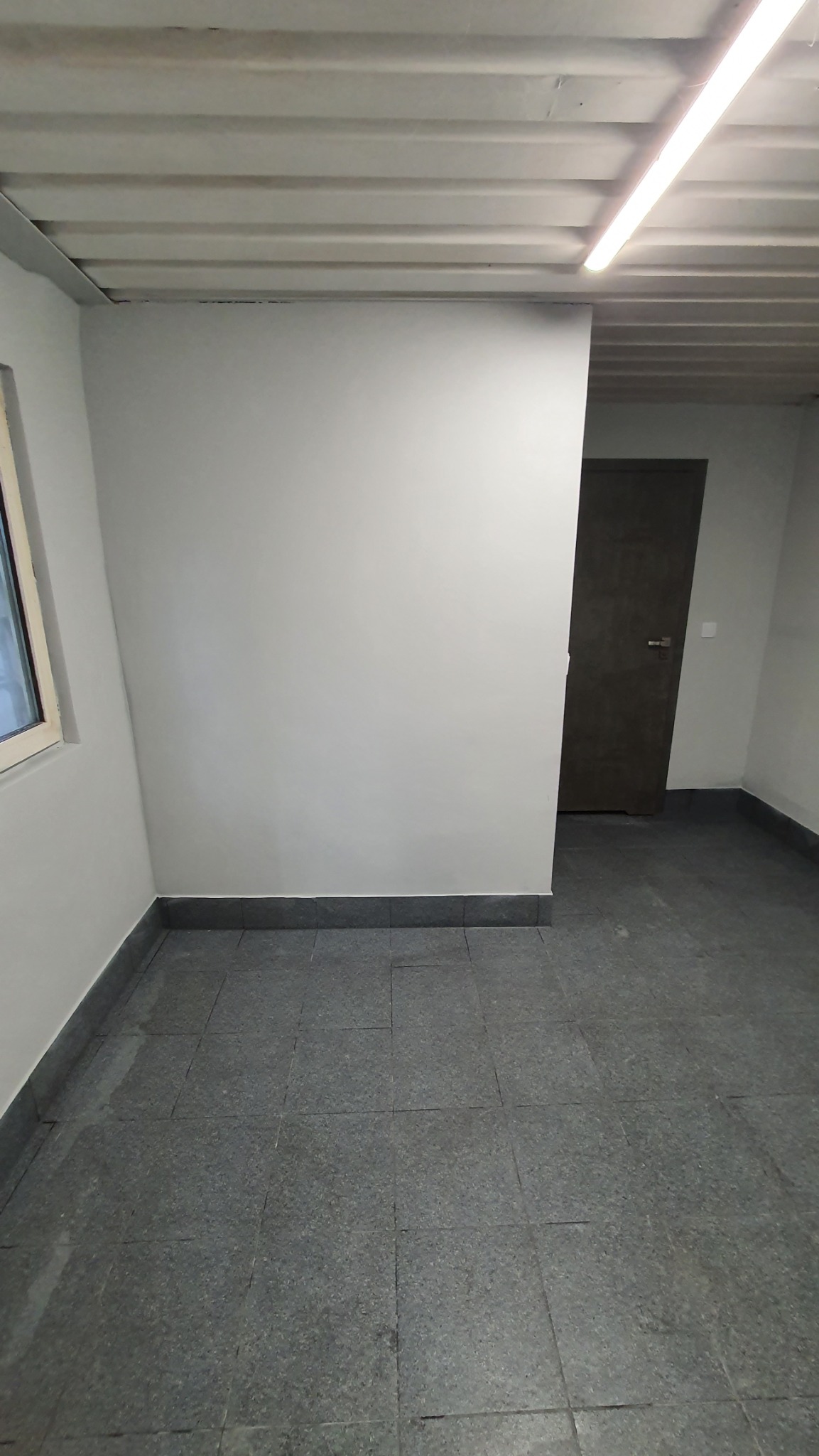 Pomalowane na szaro wnętrze pomieszczenia z szarą podłogą z płytek, widoczne okno i drzwi, sufit z widocznymi elementami konstrukcyjnymi i lampą jarzeniową.