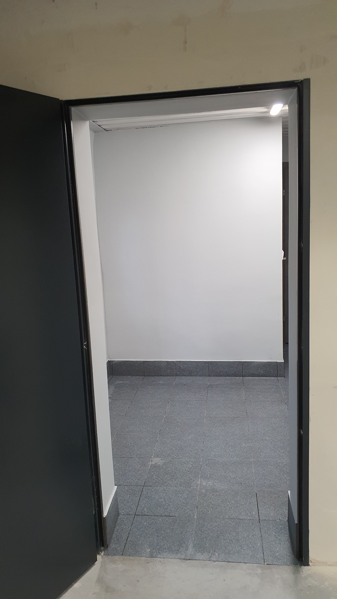 Widok przez otwarte, ciemne drzwi na wykończone wnętrze z szarą podłogą z płytek i białą ścianą, widoczny fragment listwy przypodłogowej.