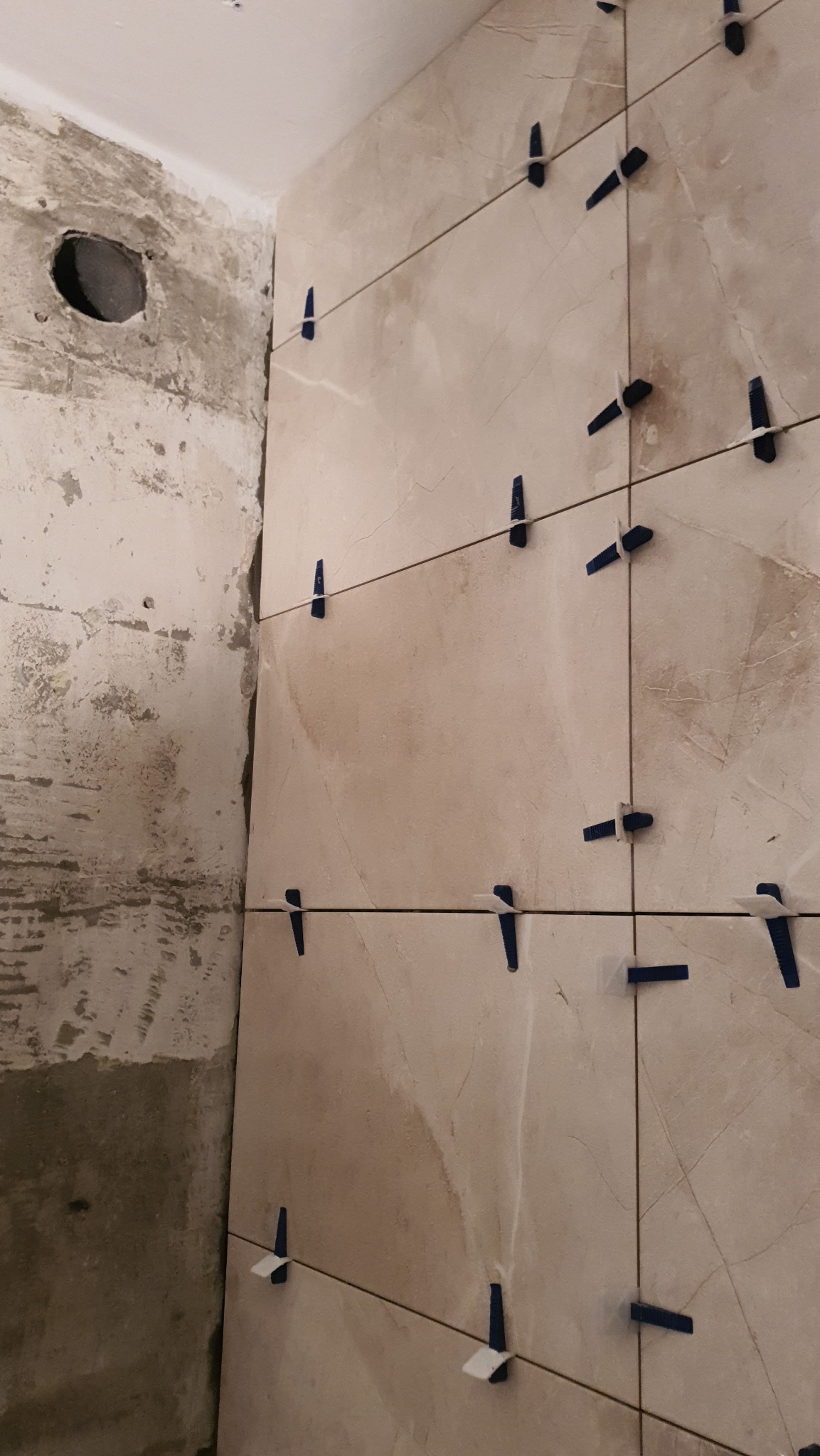 Ściana w trakcie układania płytek imitujących marmur, z użyciem niebieskich krzyżyków dystansowych, widoczny surowy fragment ściany z otworem