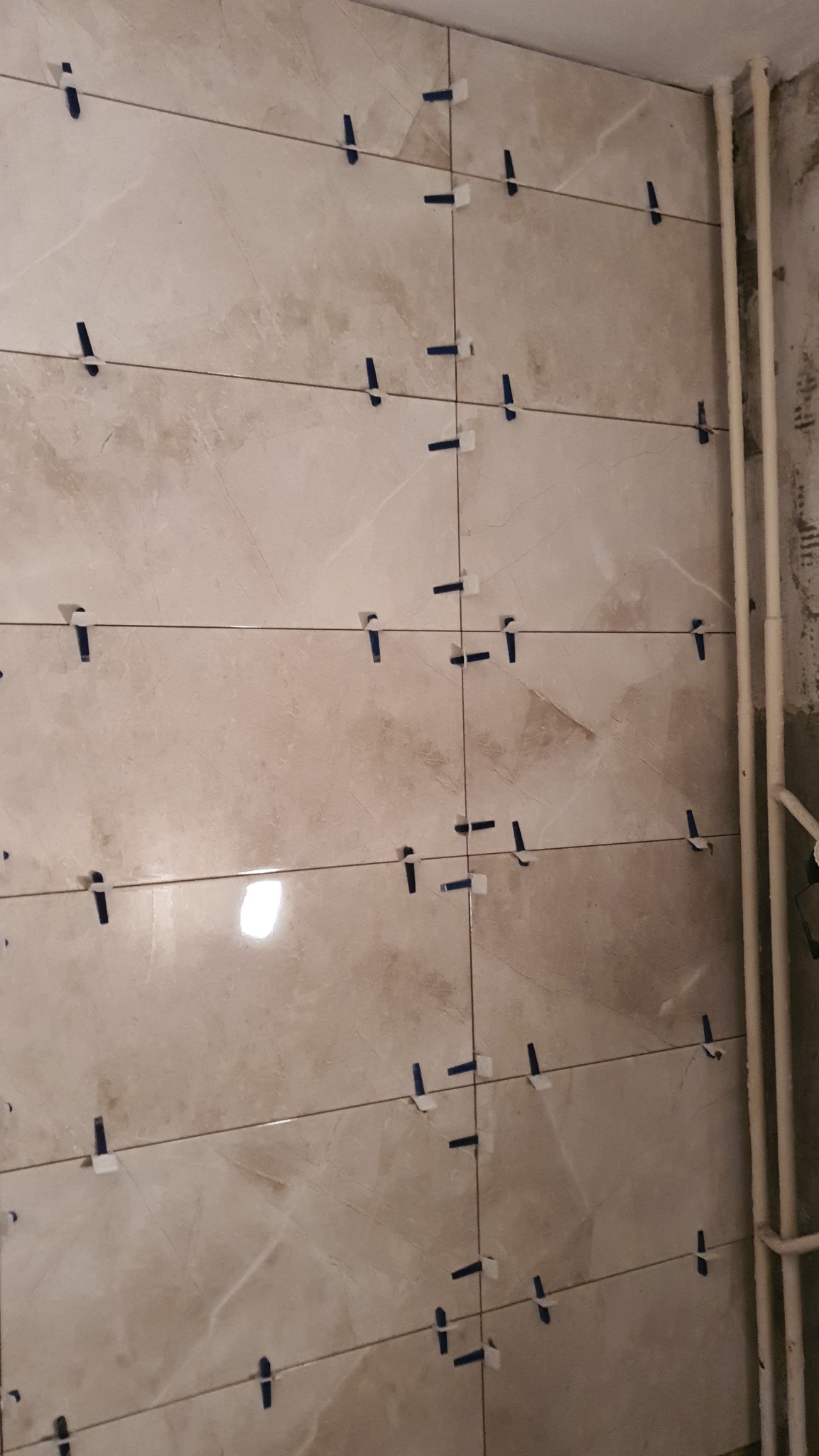 Ściana z beżowych płytek imitujących marmur, w trakcie układania, z widocznymi krzyżykami dystansowymi, rury grzewcze w prawym górnym rogu.