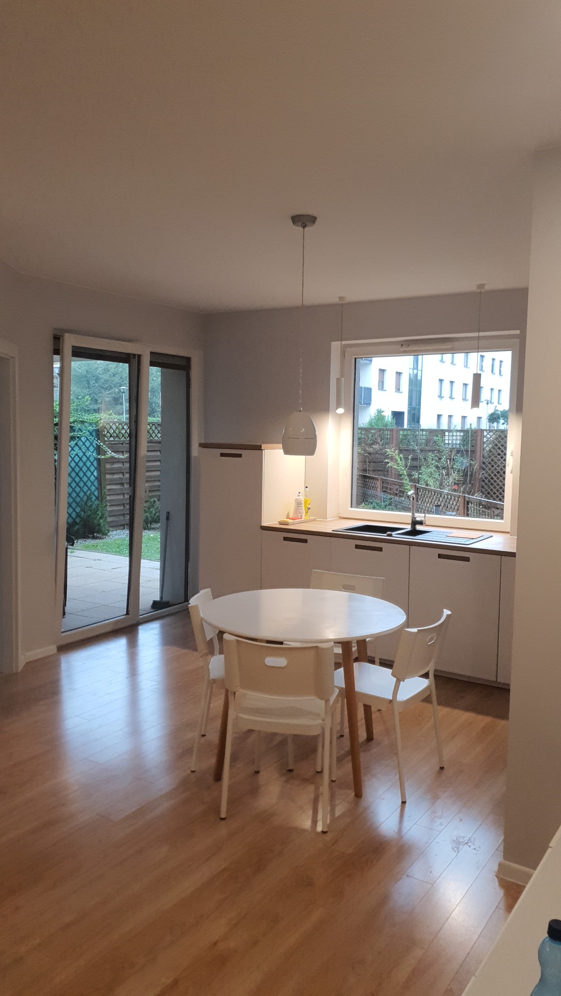 Jasne wnętrze mieszkania z drewnianą podłogą, okrągłym białym stołem i czterema krzesłami, kuchnia z białymi szafkami i drewnianym blatem pod oknem, widok na ogród przez otwarte drzwi balkonowe.