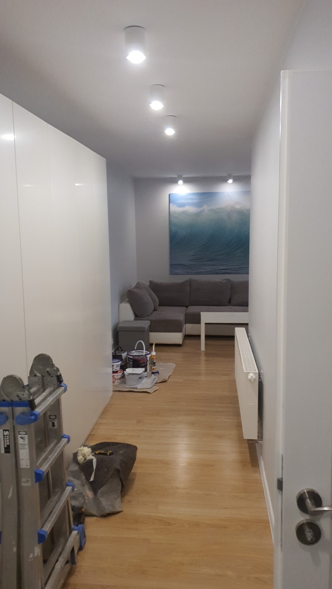 Korytarz w trakcie odświeżania: widoczne puszki z farbą, drabina malarska i zabezpieczona folią podłoga, w tle sofa i obraz z motywem fal.